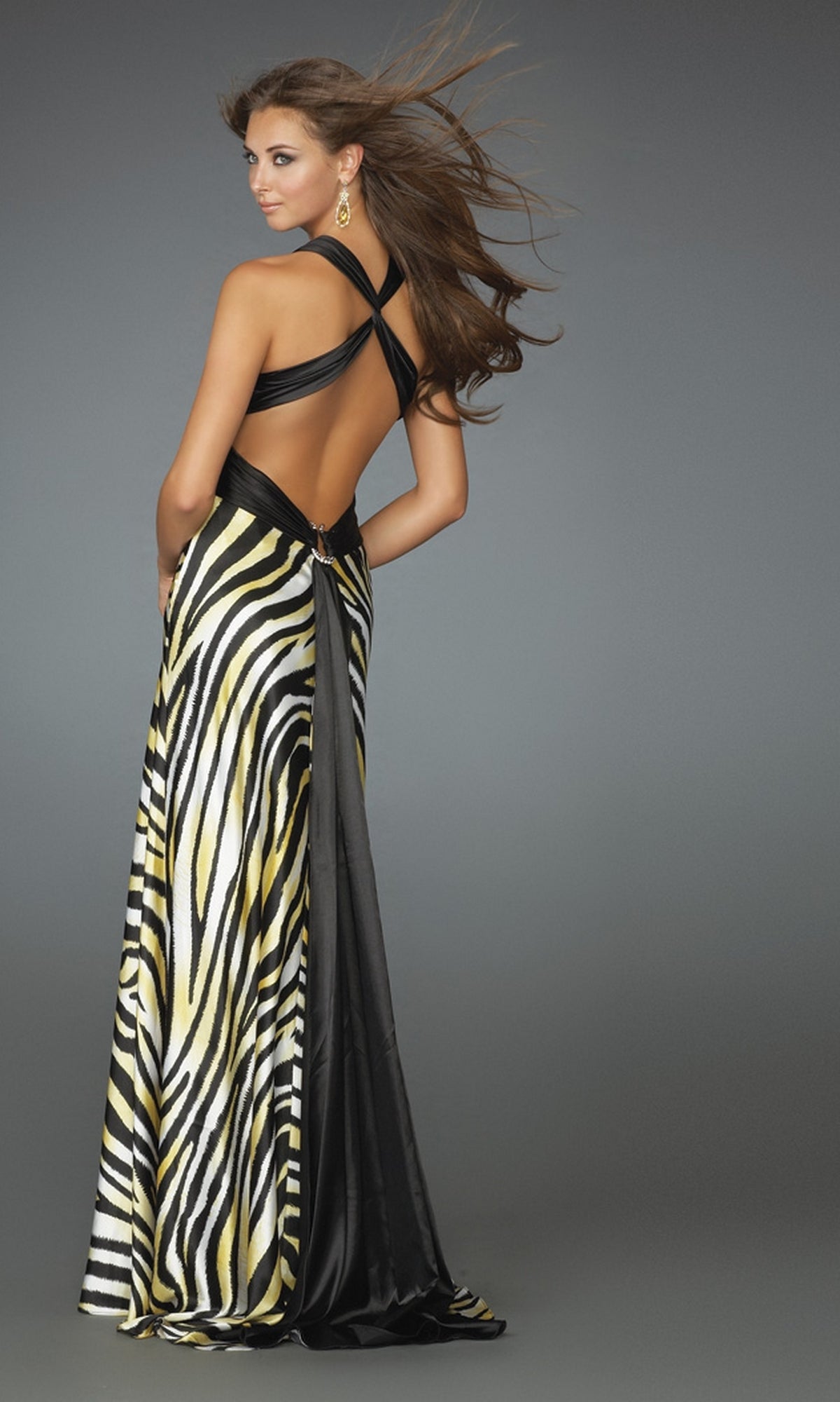 A woman poses in the La Femme Dress 14588, a striking zebra print dress with black and yellow accents and an open back, looking over her shoulder against a plain gray background.